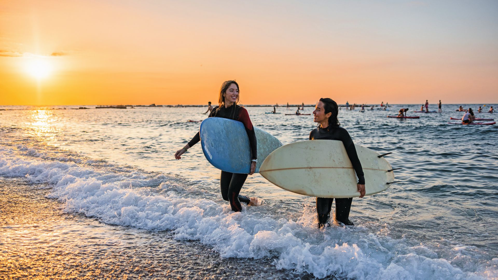 Surfers sonriendo mientras salen del agua de la playa en una puesta de sol, con mesa de surf y neopreno.