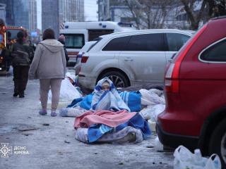 Una mujer sentada en el suelo cubierta con mantas frente a un edificio destruido por un ataque ruso.