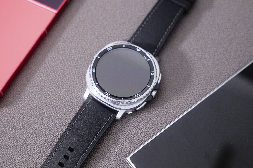Reloj inteligente con esfera redonda plateada, bisel numerado y correa de cuero negra sobre una superficie gris junto al smartphone.