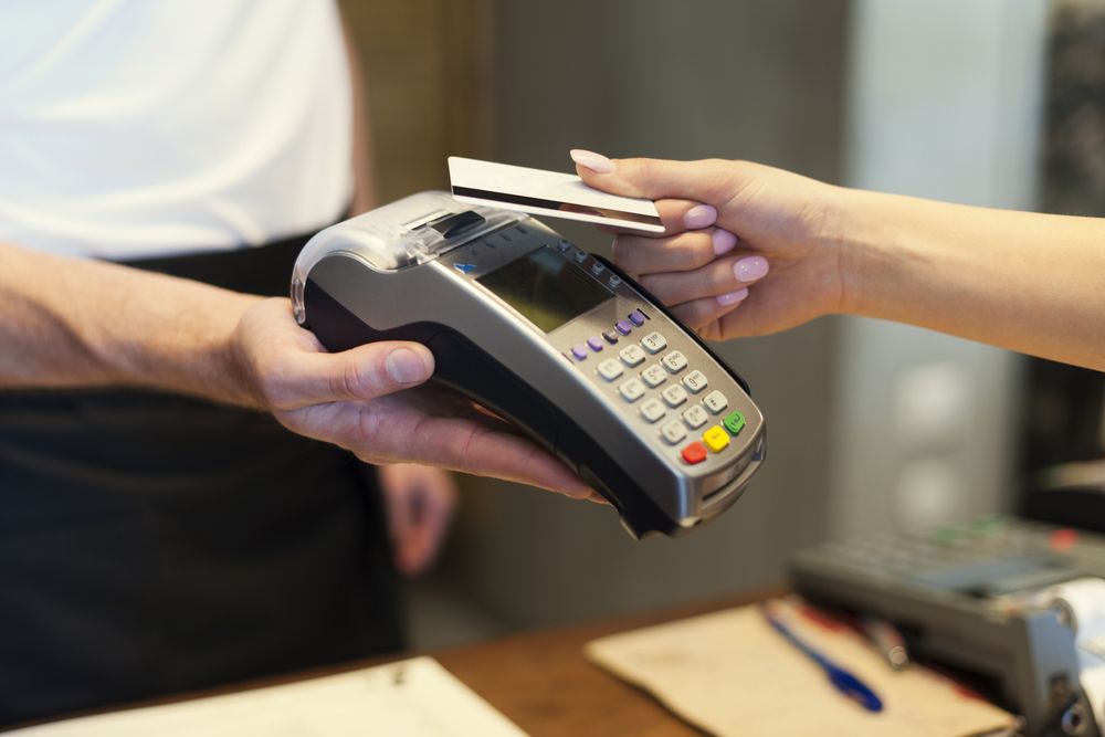 Primer plano: una persona paga con una tarjeta bancaria utilizando tecnología sin contacto en una terminal POS sostenida por un empleado de la tienda.